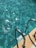 Sunlight reflects off the clear blue water in a swimming pool, highlighting ripples and waves. A metal pool ladder with two handles descends into the water, positioned along the pool edge. The surrounding area is made of a light-colored concrete material.