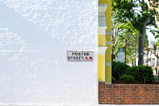 A white brick wall features a street sign that reads 'FRISTON STREET S.W.', while a portion of a yellow building and a brick wall appear alongside it. In the background, there are lush green trees and some street signs visible.