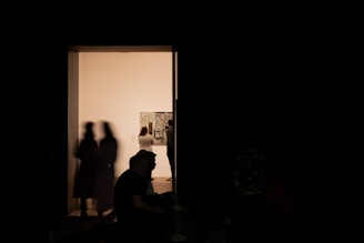 A dimly lit interior with silhouetted figures near an illuminated gallery space. The background shows a bright room with people examining artworks on the walls.
