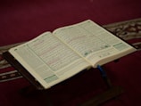 An open book rests on a wooden stand, displaying text in Arabic script. The setting is a warm, softly lit space with a maroon carpet. Intricate patterns are present on the book's pages.