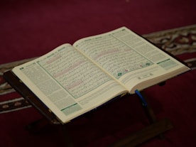 A peaceful scene of a golden Qur’an open on a wooden stand with soft sunlight streaming through a window.