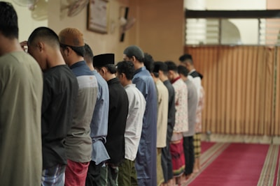 A group of men standing in a row, wearing a variety of traditional clothing including shirts, tunics, and head coverings, inside a room with muted lighting. They are lined up on a red carpet with wooden paneling in the background.