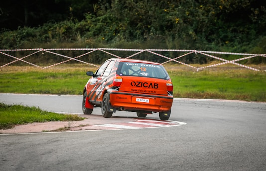 A bright orange rally car is maneuvering around a curved section of a racetrack. The car appears to be in motion with one of its rear wheels slightly off the ground, suggesting a high-speed turn. The track is bordered by grass, and there's a taped-off area with trees in the background, indicating a racing event setting.