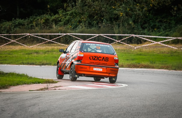 A bright orange rally car is maneuvering around a curved section of a racetrack. The car appears to be in motion with one of its rear wheels slightly off the ground, suggesting a high-speed turn. The track is bordered by grass, and there's a taped-off area with trees in the background, indicating a racing event setting.