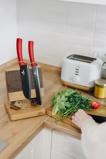 A small kitchen counter with wooden surfaces featuring a knife block with two knives that have red handles. Nearby, there is a person’s hand cutting fresh herbs and vegetables on a cutting board. A white toaster and a jar of spices are also present on the counter.