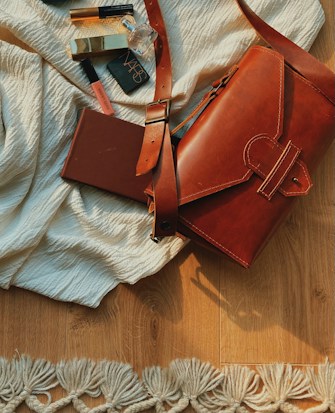 On a wooden floor, a brown leather bag rests partially open, surrounded by various cosmetic products including a lipstick and a perfume bottle, along with a closed brown notebook. The setup is laid on a crumpled white fabric with a textured, decorative fringe.