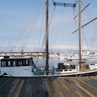 A wooden dock leads to an old-fashioned sailing ship with tall masts and numerous ropes. The ship's hull shows signs of age and wear. In the background, a marina is filled with modern sailboats and yachts, set against a clear blue sky.