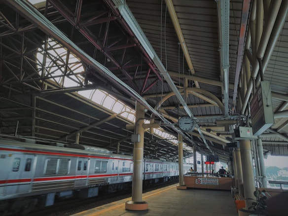 A train platform with a train in motion, covered by a high, industrial-style metal roof with exposed beams and skylights. The platform has signs, a digital display, and a wall clock showing the time as noon or midnight. The environment looks utilitarian, with columns supporting the roof and various cables and piping visible.