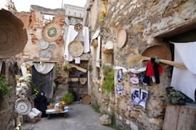 A rustic alleyway with stone walls decorated with woven baskets and various traditional crafts. Colorful textiles and images hang on the walls, alongside dried plants and vibrant flowers. The atmosphere is quaint and artistic, with elements of a rural life displayed throughout the setting.