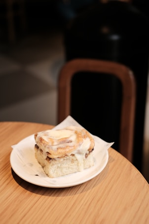A freshly baked cinnamon roll covered in a generous layer of creamy icing is placed on a white plate, set on a wooden table. The background is slightly blurred, featuring a portion of a dark chair and softly lit flooring.