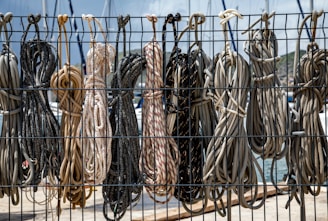 Several coils of rope hang from a metal fence, neatly arranged in a row. The ropes vary in color and texture, with some displaying striped patterns. In the background, elements of a marina are visible, including parts of sailboats and a glimpse of the water.