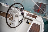 A yacht cockpit featuring a large steering wheel with a metal and rubber grip, surrounded by wooden slat seating and floors. A winch with a rope is visible to the side, alongside various nautical controls. The layout is clean and organized, with ropes neatly coiled and stowed.
