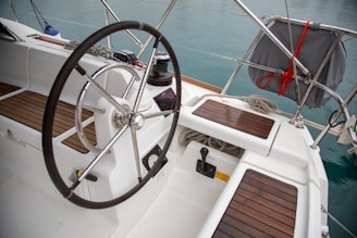 A yacht cockpit featuring a large steering wheel with a metal and rubber grip, surrounded by wooden slat seating and floors. A winch with a rope is visible to the side, alongside various nautical controls. The layout is clean and organized, with ropes neatly coiled and stowed.