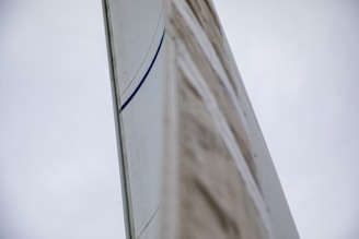 A close-up view of a sail with a visible blue stripe against a cloudy sky backdrop. The perspective emphasizes the height and texture of the sail.