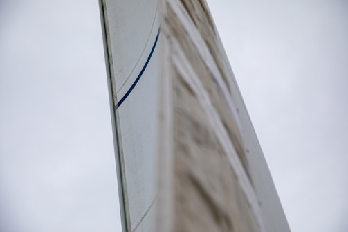 A close-up view of a sail with a visible blue stripe against a cloudy sky backdrop. The perspective emphasizes the height and texture of the sail.