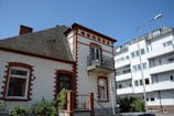 A traditional two-story house with red brick accents and a small balcony is positioned next to a more modern apartment building. The house features a sloped roof with a small attic window. A streetlamp and some greenery, including shrubs and vines, are present in the foreground.