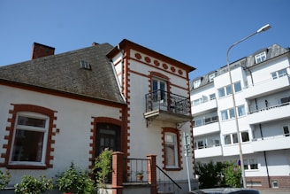 A traditional two-story house with red brick accents and a small balcony is positioned next to a more modern apartment building. The house features a sloped roof with a small attic window. A streetlamp and some greenery, including shrubs and vines, are present in the foreground.