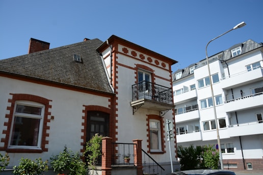 A traditional two-story house with red brick accents and a small balcony is positioned next to a more modern apartment building. The house features a sloped roof with a small attic window. A streetlamp and some greenery, including shrubs and vines, are present in the foreground.