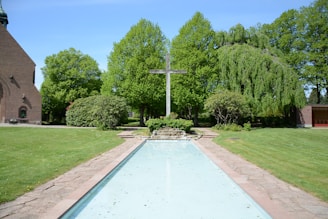 A serene outdoor scene featuring a cross at the center, surrounded by lush green trees and bushes. A pathway and a shallow, reflective water feature are in the foreground. A brick building is partially visible to the left, adding to the peaceful garden setting.