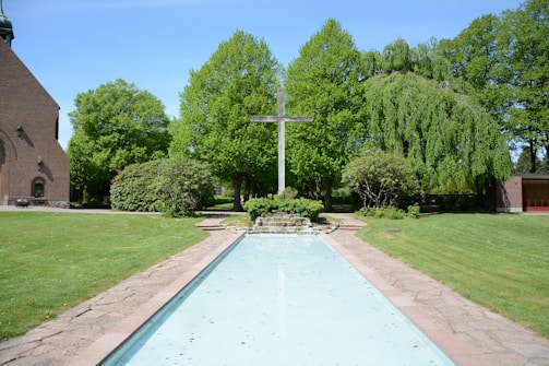 A serene outdoor scene featuring a cross at the center, surrounded by lush green trees and bushes. A pathway and a shallow, reflective water feature are in the foreground. A brick building is partially visible to the left, adding to the peaceful garden setting.