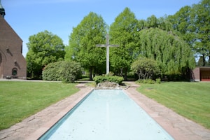 A serene outdoor scene featuring a cross at the center, surrounded by lush green trees and bushes. A pathway and a shallow, reflective water feature are in the foreground. A brick building is partially visible to the left, adding to the peaceful garden setting.