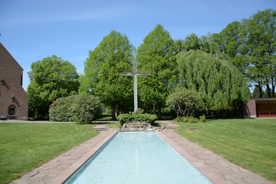 A serene outdoor setting featuring a large wooden cross centrally positioned in a landscaped area. The scene includes a reflecting pool leading towards the cross, surrounded by well-maintained greenery and trees. There is a brick building to the left and another structure partially visible on the right.