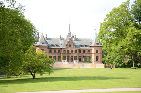 A large, historic brick mansion with pointed towers and a steep roof is surrounded by lush greenery. The lawn in front is expansive, with well-maintained grass. There are tall trees on either side of the building, providing a picturesque and tranquil setting.