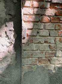 A weathered brick wall partially covered in rough, textured plaster. The bricks are various shades of red and brown, while the plaster is gray and unevenly applied. Light and shadows create a contrast on the surface.