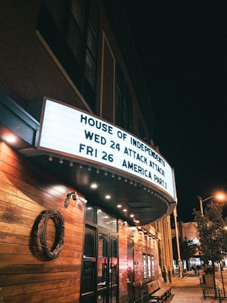 A street view at night with a large theater marquee displaying upcoming show dates and names, set against a building with wooden paneling and decorative lighting. Several small trees and streetlights are visible along the sidewalk, creating an inviting atmosphere.