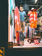 A store display featuring two mannequins dressed in tropical-themed summer attire. One mannequin wears a blue Hawaiian shirt and khaki shorts with a straw hat, while the other is in a red and orange outfit with a similar hat. In front of them, two skateboards are placed upright. The background shows racks of clothing, and there is a sign indicating the store is an authorized dealer.