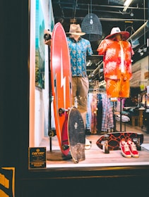 A store display featuring two mannequins dressed in tropical-themed summer attire. One mannequin wears a blue Hawaiian shirt and khaki shorts with a straw hat, while the other is in a red and orange outfit with a similar hat. In front of them, two skateboards are placed upright. The background shows racks of clothing, and there is a sign indicating the store is an authorized dealer.
