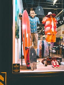 A store display featuring two mannequins dressed in tropical-themed summer attire. One mannequin wears a blue Hawaiian shirt and khaki shorts with a straw hat, while the other is in a red and orange outfit with a similar hat. In front of them, two skateboards are placed upright. The background shows racks of clothing, and there is a sign indicating the store is an authorized dealer.