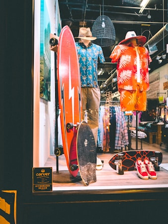 A store display featuring two mannequins dressed in tropical-themed summer attire. One mannequin wears a blue Hawaiian shirt and khaki shorts with a straw hat, while the other is in a red and orange outfit with a similar hat. In front of them, two skateboards are placed upright. The background shows racks of clothing, and there is a sign indicating the store is an authorized dealer.