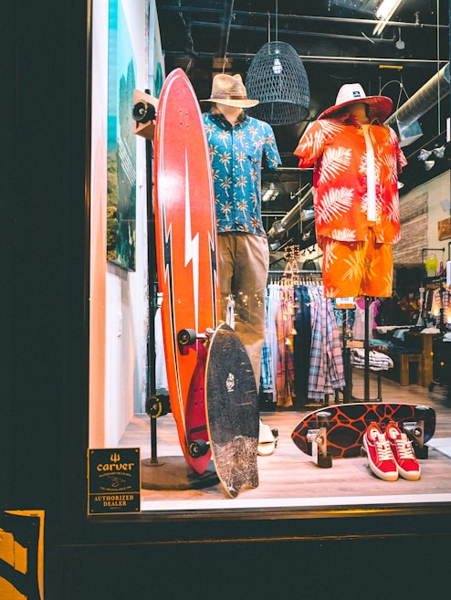 A store display featuring two mannequins dressed in tropical-themed summer attire. One mannequin wears a blue Hawaiian shirt and khaki shorts with a straw hat, while the other is in a red and orange outfit with a similar hat. In front of them, two skateboards are placed upright. The background shows racks of clothing, and there is a sign indicating the store is an authorized dealer.
