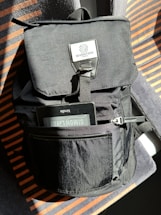 A black fabric backpack with a flap and front pocket is placed on a seat with orange and grey striped upholstery. An e-reader device is partially visible in the pocket, displaying the name 'Simon Leay' on the screen.