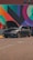 A dark gray car with its hood open is parked in front of a vibrant mural featuring geometric shapes and a variety of colors including red, orange, blue, and green. Two people are standing nearby, possibly inspecting or discussing the car.