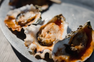 A close-up view of several oysters on a metal plate, with the shells partially open and the inner edible part visible. The oysters are garnished with a red sauce, which adds a vibrant contrast to the natural tones of the shells. The lighting casts soft shadows, highlighting the texture of the shells and the surface of the sauce.