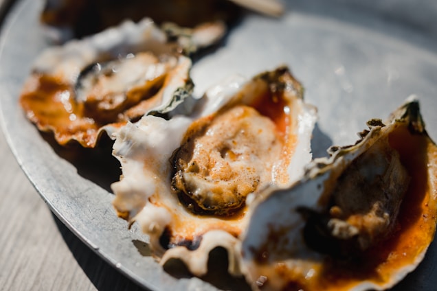 A close-up view of several oysters on a metal plate, with the shells partially open and the inner edible part visible. The oysters are garnished with a red sauce, which adds a vibrant contrast to the natural tones of the shells. The lighting casts soft shadows, highlighting the texture of the shells and the surface of the sauce.