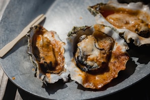 Three oysters on half shells are served on a metal plate, drizzled with a spicy or tangy sauce. A small wooden fork lies nearby, and the lighting creates shadows, emphasizing the texture of the oysters and sauce.