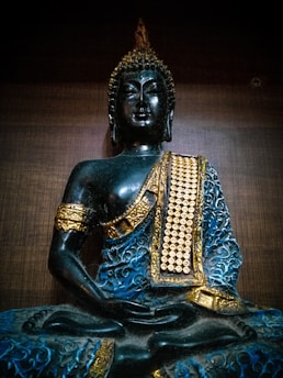 A black statue of Buddha adorned with golden and blue decorative elements, sitting in a meditative pose against a dark wooden background.