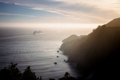 A large cruise ship sails towards the horizon on a calm sea, with a rugged coastline on the right. The setting or rising sun casts a golden glow across the water and cliffs, creating a peaceful atmosphere. Trees are visible on the cliff edge, silhouetted against the soft light.