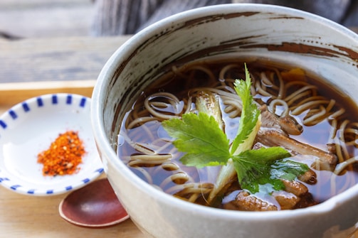 A bowl of broth with noodles garnished with fresh greens and mushrooms. A side plate with red spices and a wooden spoon are nearby, set on a wooden surface.