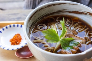 A bowl of broth with noodles garnished with fresh greens and mushrooms. A side plate with red spices and a wooden spoon are nearby, set on a wooden surface.