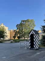 A small striped guardhouse stands on the side of a spacious, paved area. Nearby, a matching striped barrier blocks part of the pathway. Behind them, a cluster of trees provides shade, and imposing yellow buildings with white trim rise in the background. The sky is a clear blue.