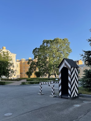 A small striped guardhouse stands on the side of a spacious, paved area. Nearby, a matching striped barrier blocks part of the pathway. Behind them, a cluster of trees provides shade, and imposing yellow buildings with white trim rise in the background. The sky is a clear blue.