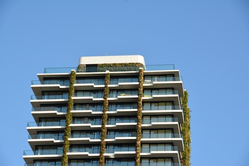 A modern high-rise building with multiple balconies is covered with vertical greenery. The building's facade is a mix of glass and white structural elements, set against a clear blue sky, creating a striking architectural design.
