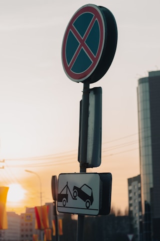 A 'no parking' sign with a towing warning depicted below, positioned against a backdrop of a modern cityscape at sunset. The sky has warm hues, and the sun casts a soft glow on the buildings.