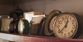 A collection of vintage clocks is arranged on a shelf, with some appearing to be old-fashioned analog clocks. The lighting creates a warm and nostalgic atmosphere, highlighting the varied designs and colors of the clocks. The focus is on a round clock with bold black numbers on a white face.