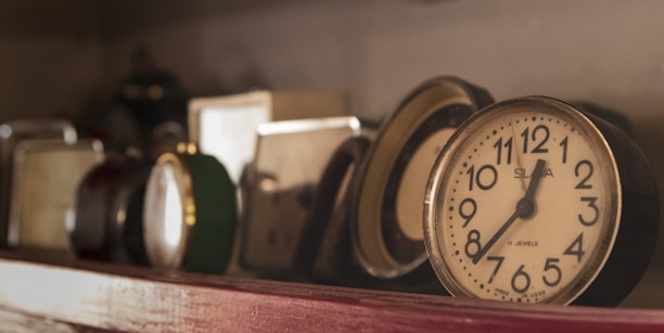 A collection of vintage clocks is arranged on a shelf, with some appearing to be old-fashioned analog clocks. The lighting creates a warm and nostalgic atmosphere, highlighting the varied designs and colors of the clocks. The focus is on a round clock with bold black numbers on a white face.