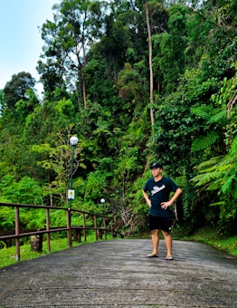 A person stands confidently on a pathway surrounded by lush green foliage and tall trees. The pathway features a wooden railing and is lined with street lamps.